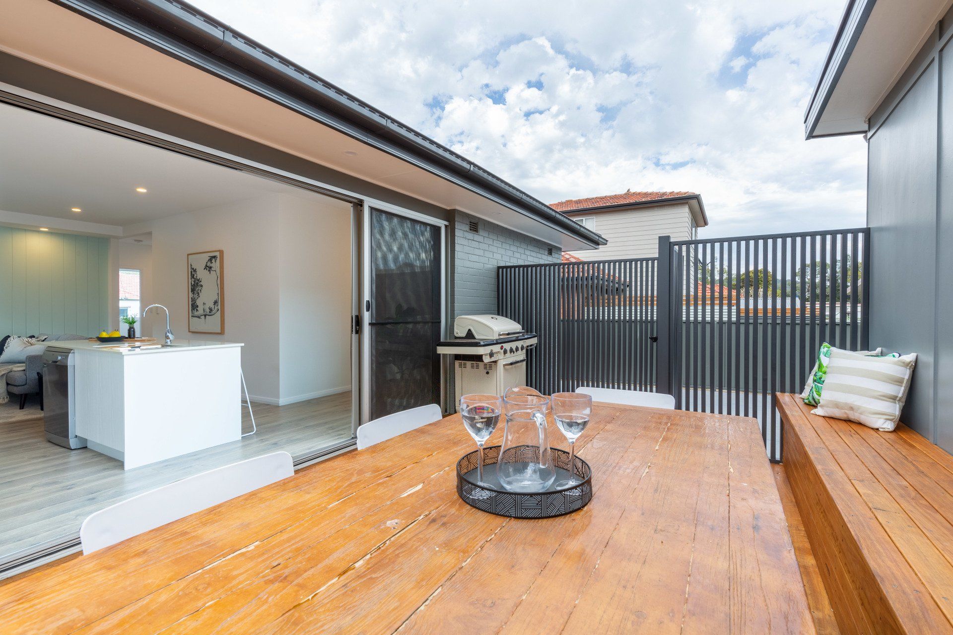Outdoor patio with wooden table, bench, and open door to a kitchen. Grill and glasses. Cloudy sky.
