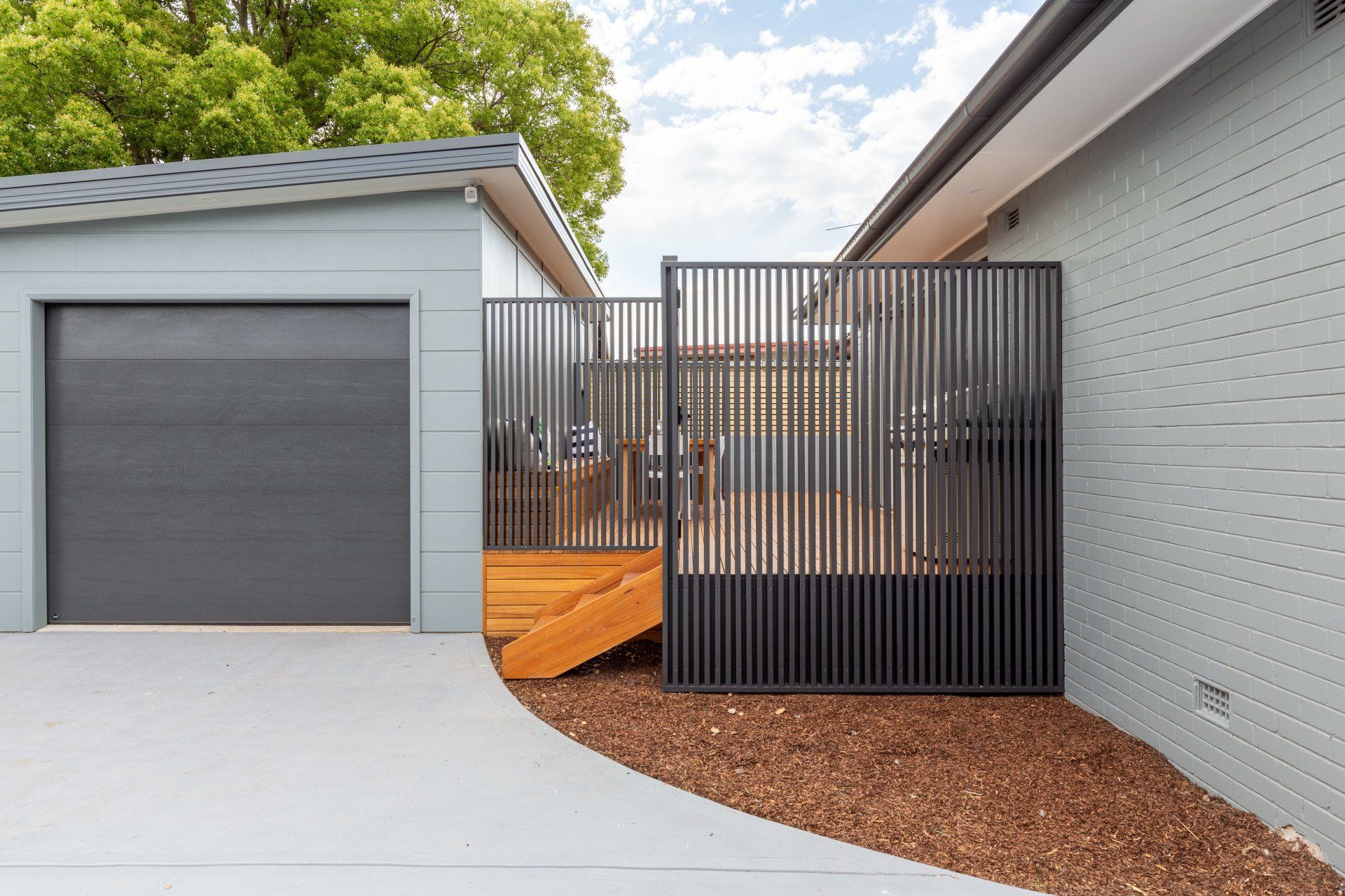 Gray garage next to a sectioned black metal gate on a wood platform, with a gray paved drive and mulch.