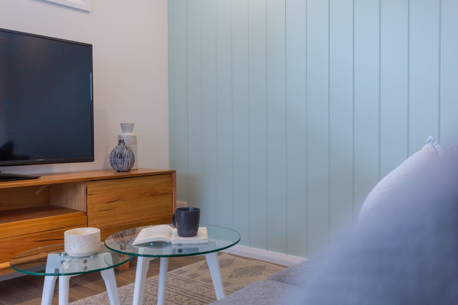 Living room corner with TV, wooden cabinet, glass tables, and light blue paneled wall.