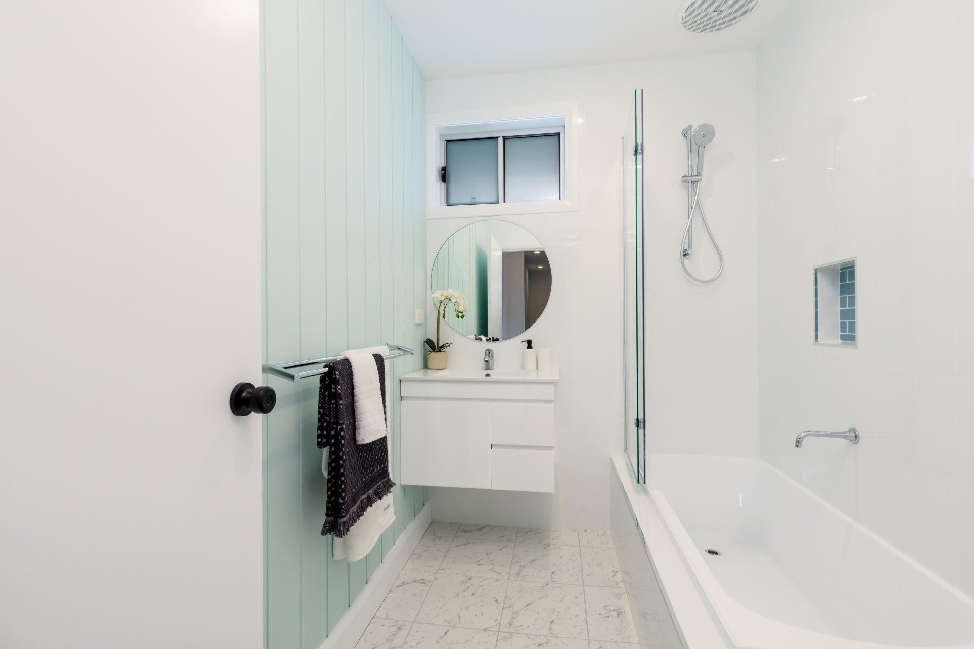 Modern bathroom: white walls, bathtub, vanity, green accent wall, round mirror, and glass shower door.