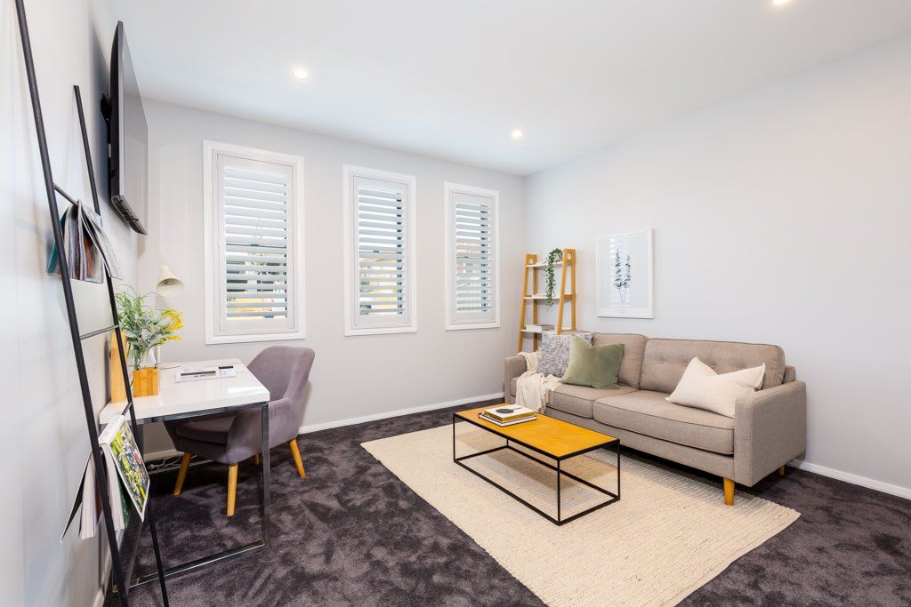 A small living room with a desk, sofa, and coffee table, all on dark carpet. Three windows on the back wall.