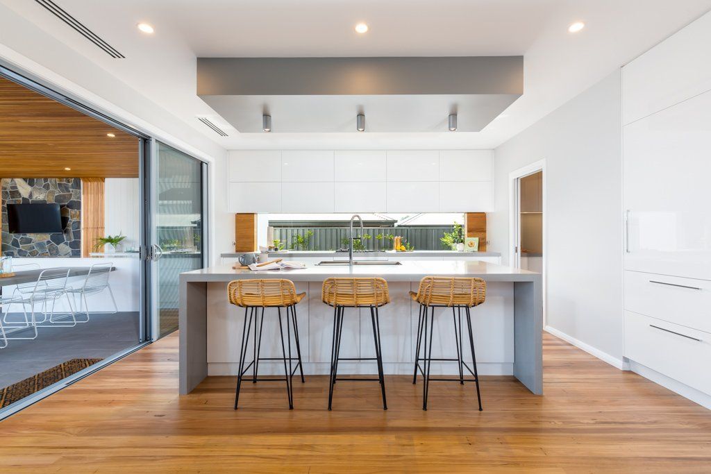 Modern white kitchen with a gray island and three bar stools. Sliding glass doors open to a living area.