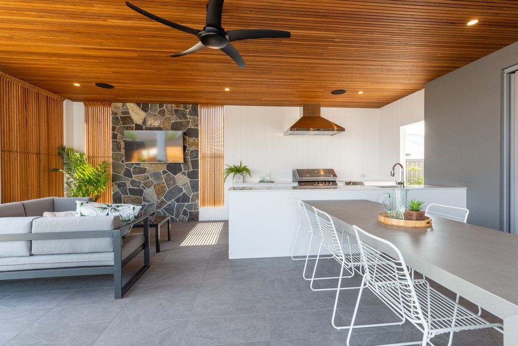 Outdoor kitchen and dining area with gray seating, white table and chairs, stone accent wall, and wood ceiling.