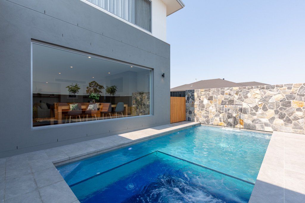 Modern home with a pool. Gray exterior, large glass window, dining table visible inside. Blue pool with stone wall backdrop.
