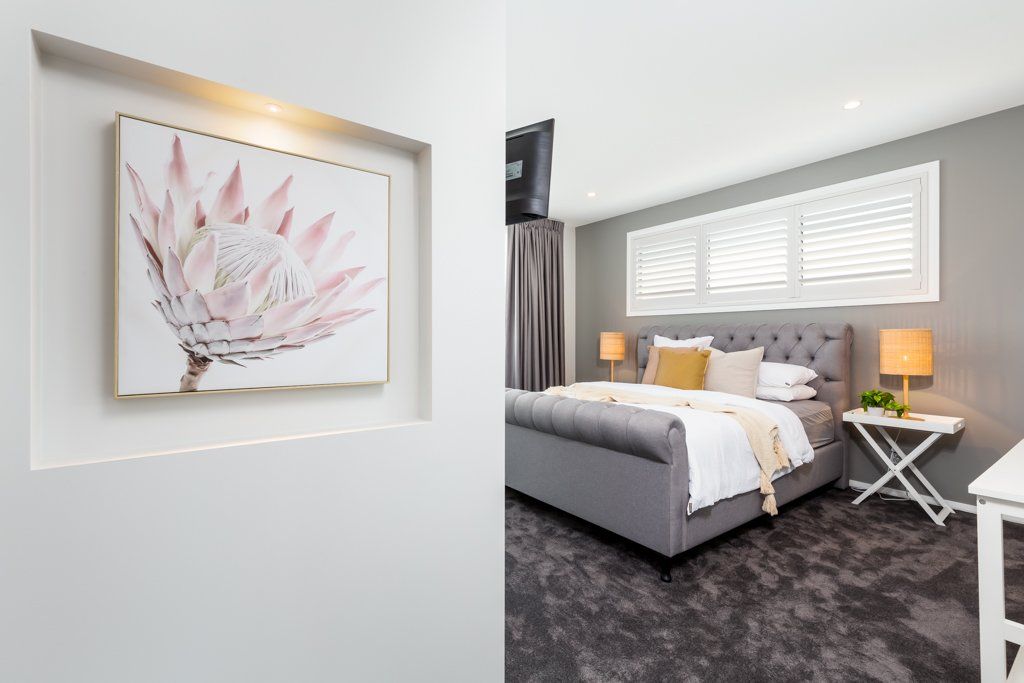 Bedroom with grey tufted bed, floral artwork in a wall niche, and a white side table.