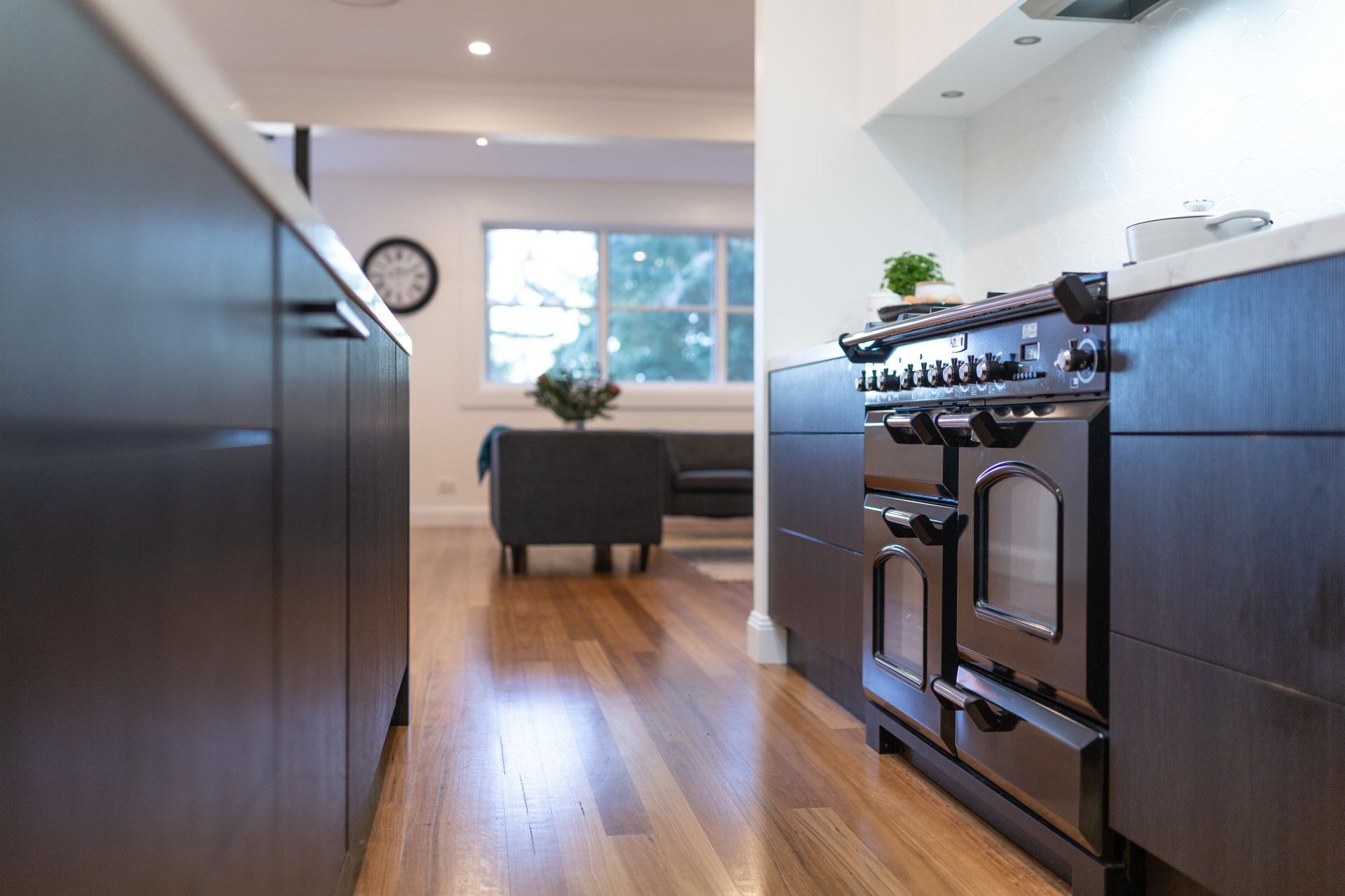 Modern kitchen with dark cabinetry, a black range, and hardwood floors leading to a living area.