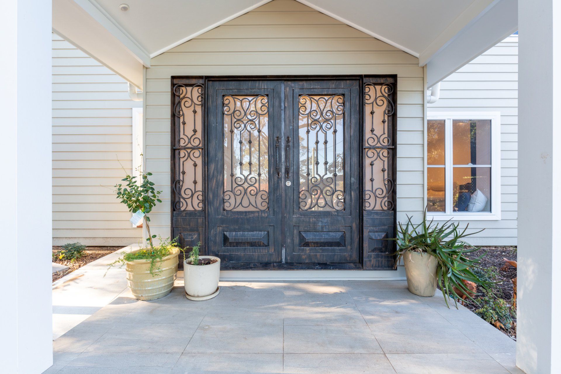 Double dark doors with decorative ironwork on a home's porch, flanked by potted plants.