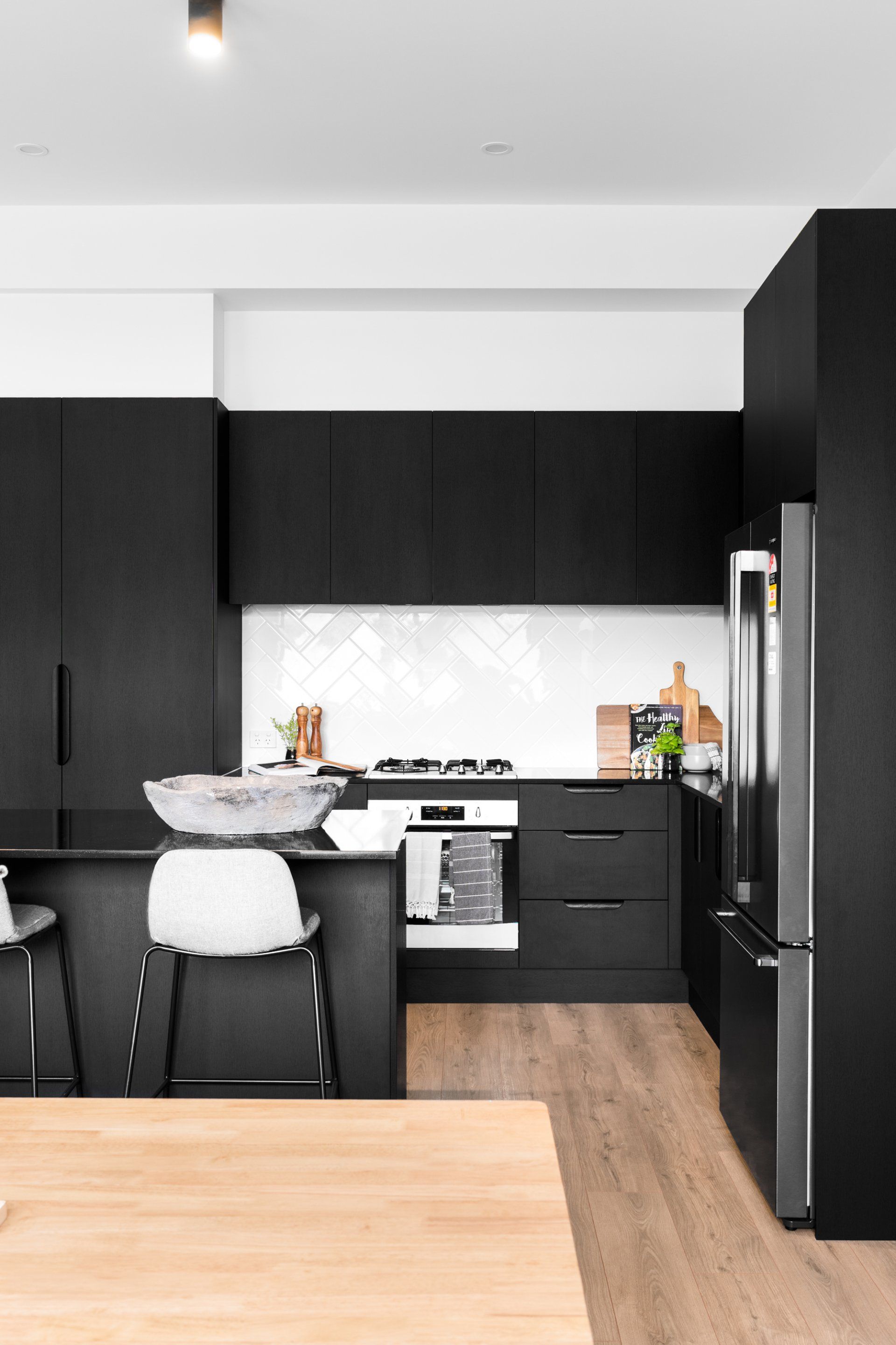 Modern black kitchen with white backsplash, stainless steel refrigerator, and wood floor.