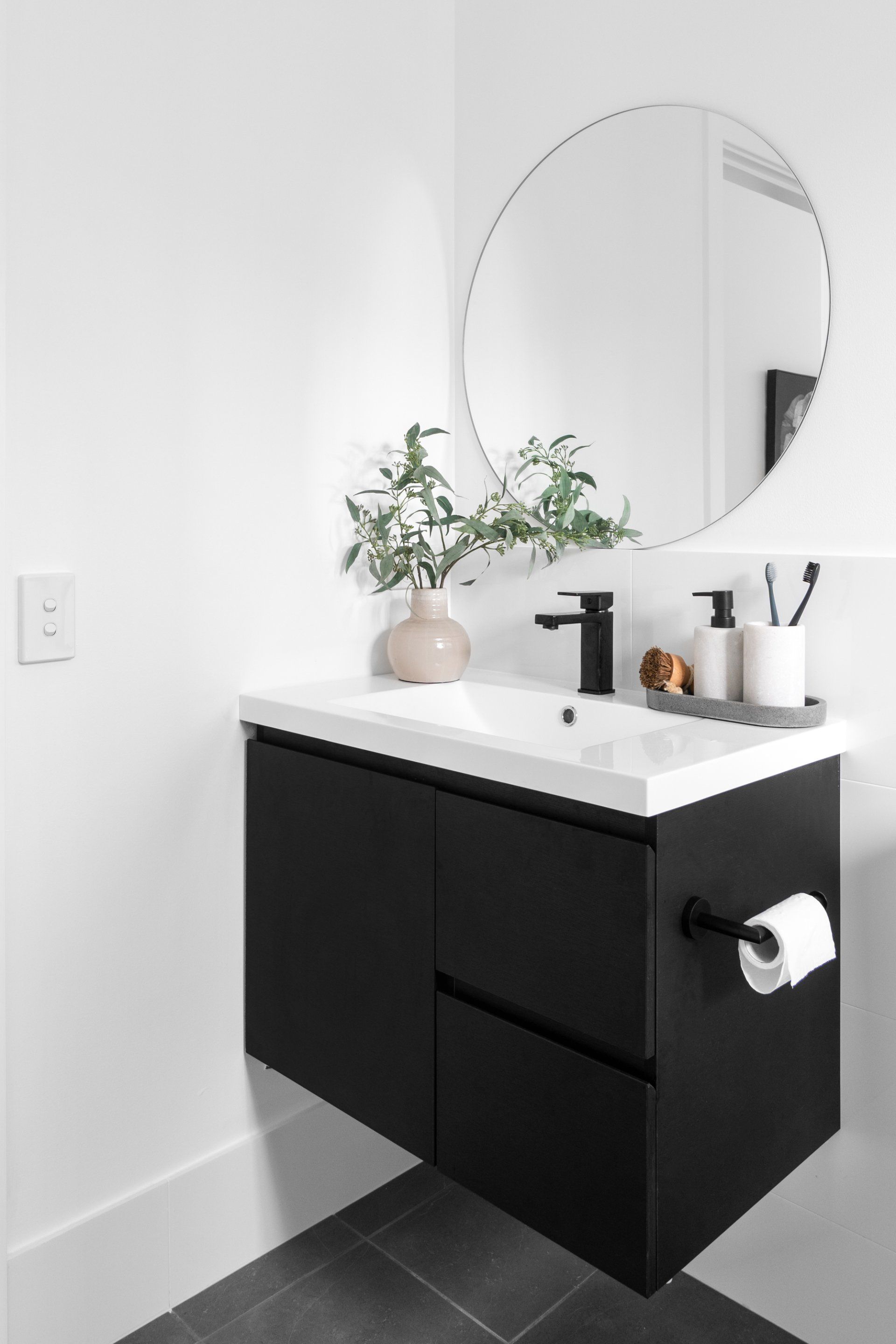 Black floating vanity with white sink, round mirror, and greenery in a minimalist bathroom.