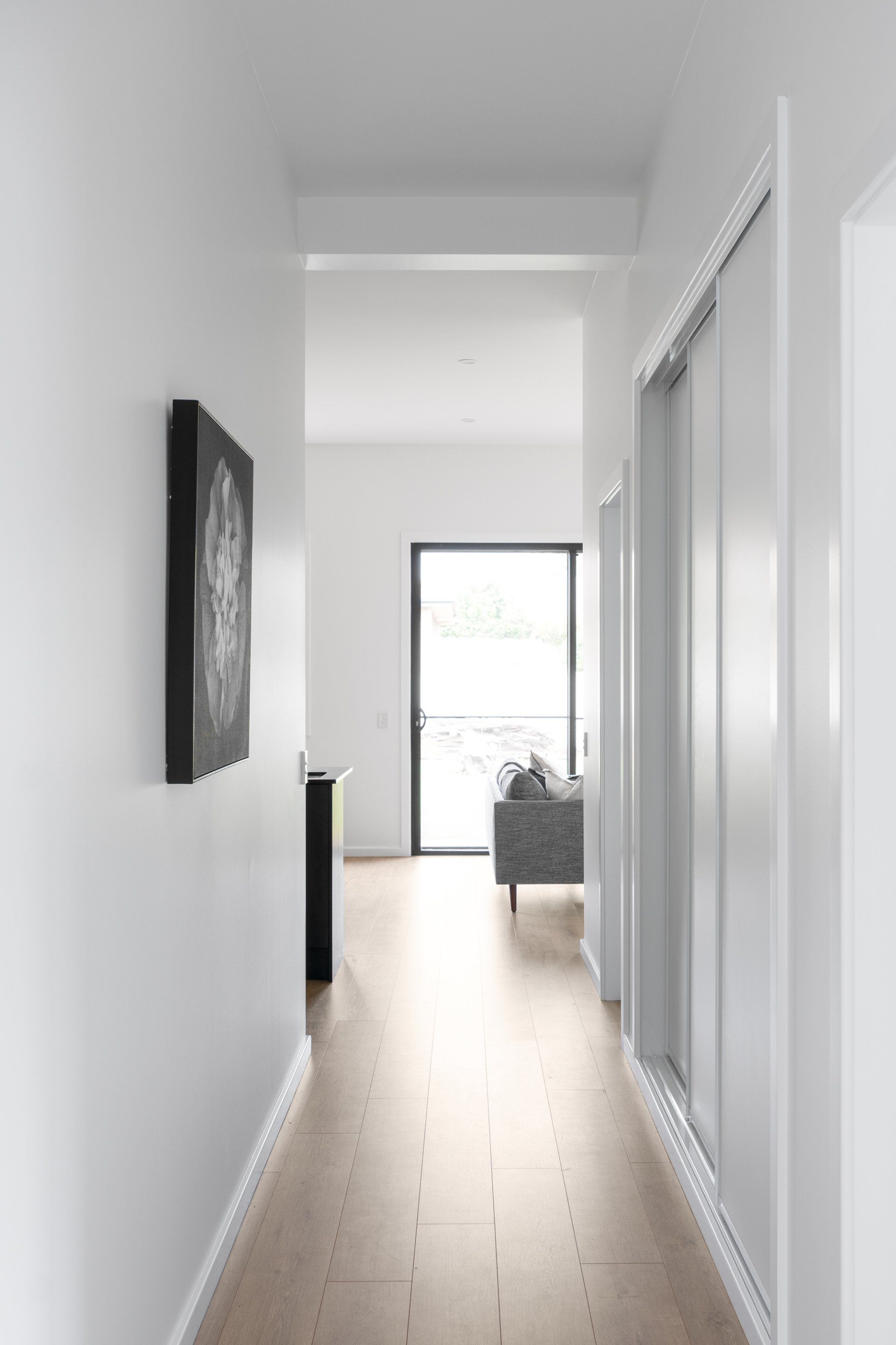 Narrow, white hallway with a light-colored wooden floor, artwork on the left wall, and a view of a living area.