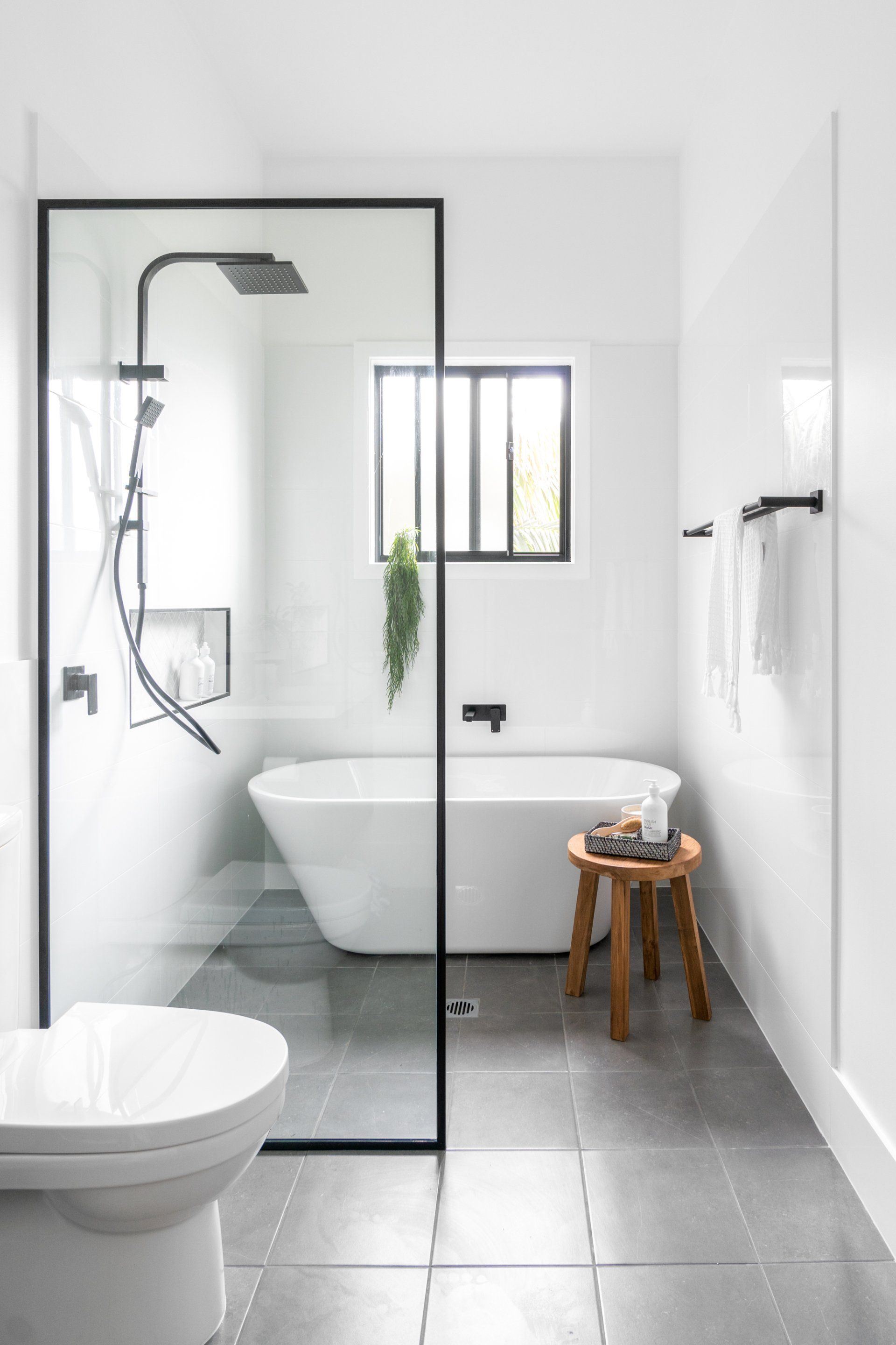 Modern white bathroom with glass shower, freestanding tub, and wooden stool.
