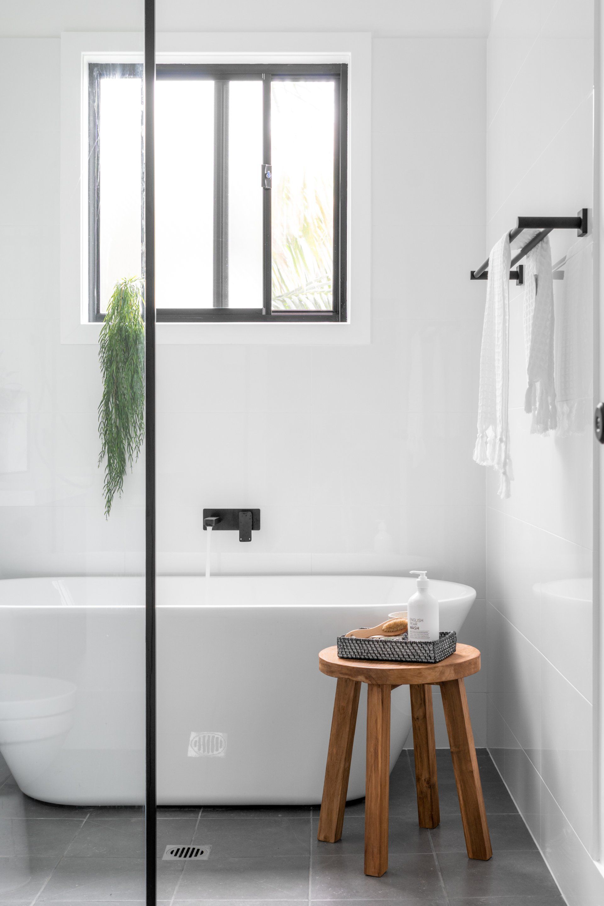 Modern white bathroom with freestanding tub, wooden stool, black fixtures, and a window.