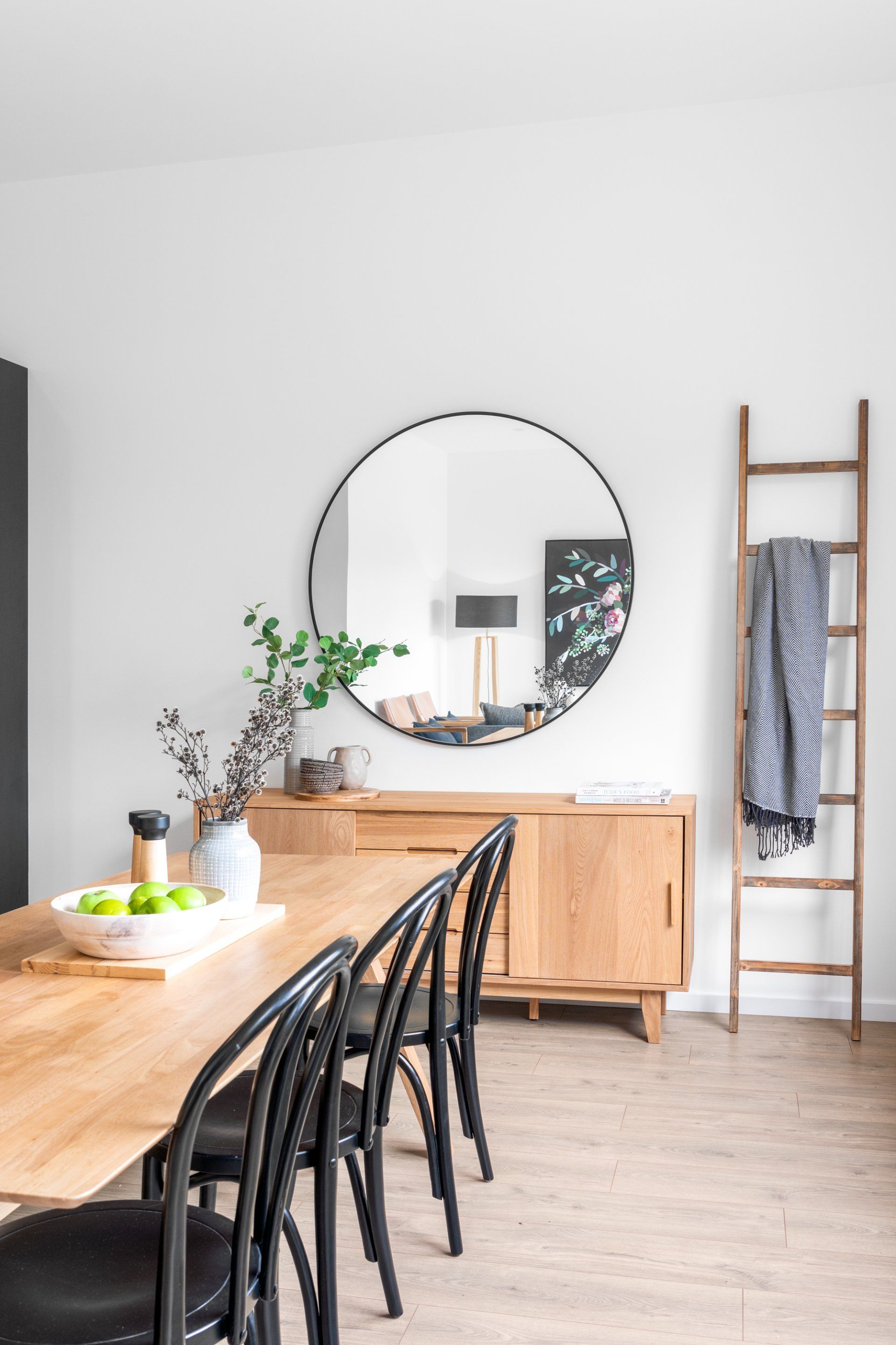 Dining room with wooden table, black chairs, buffet, round mirror, and ladder.