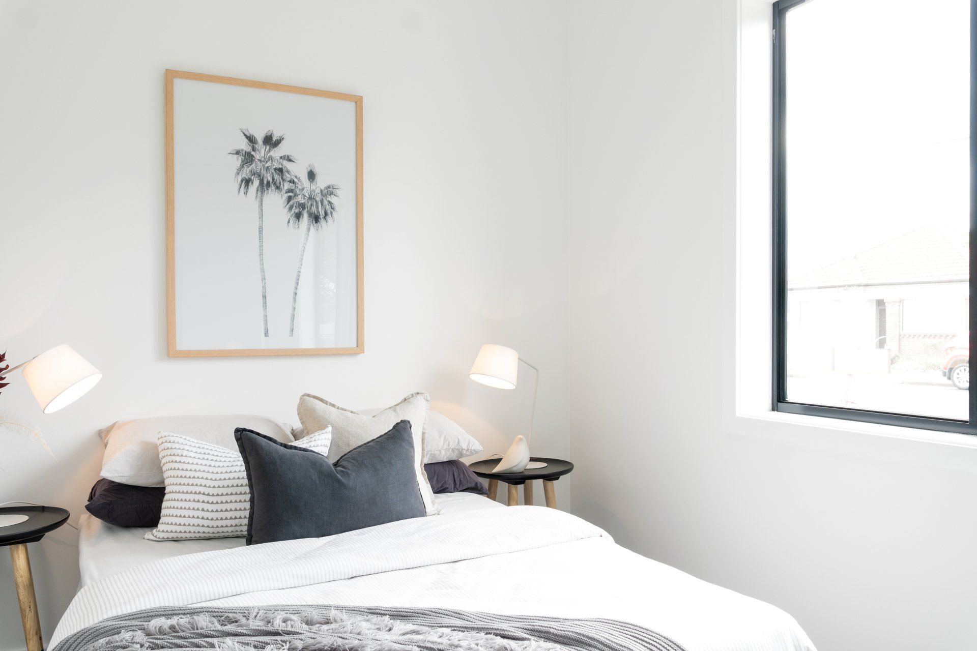 Bedroom with white walls, bed with gray pillows, palm tree artwork, and black-framed window.