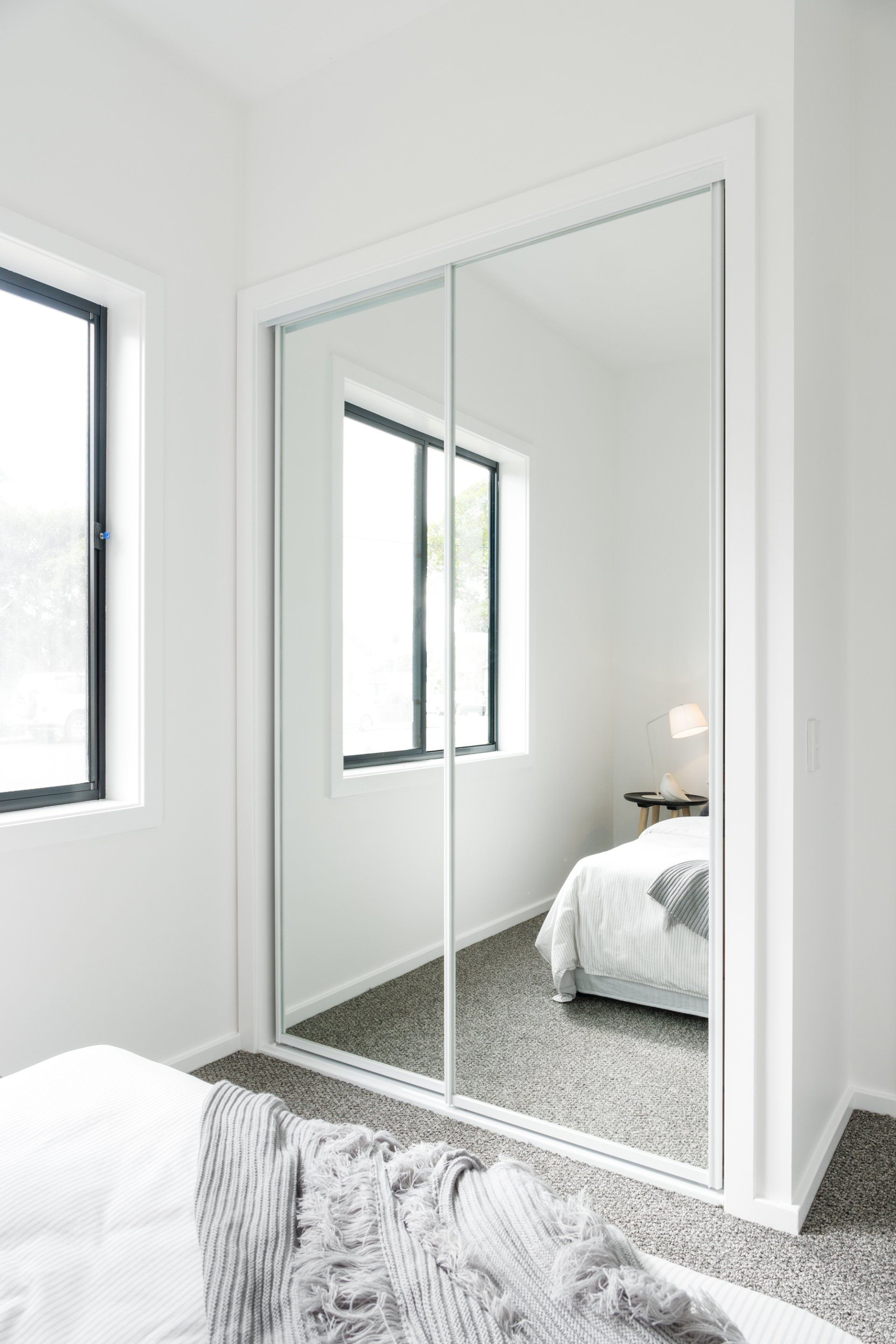 Mirrored closet doors reflect a white bedroom with bed, windows, and rug.