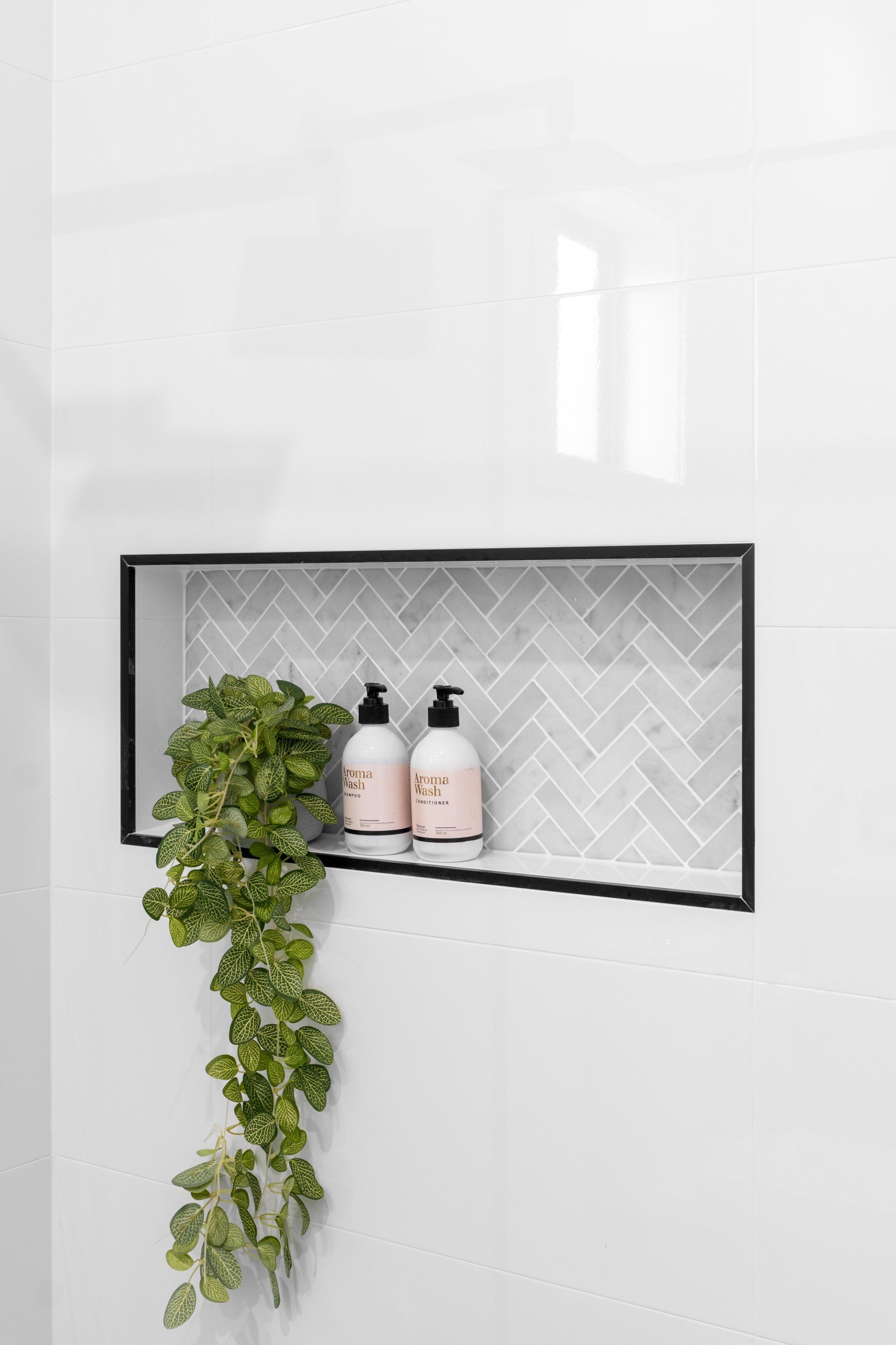 Built-in shower niche with herringbone tile, black trim, soap bottles, and a trailing green plant.