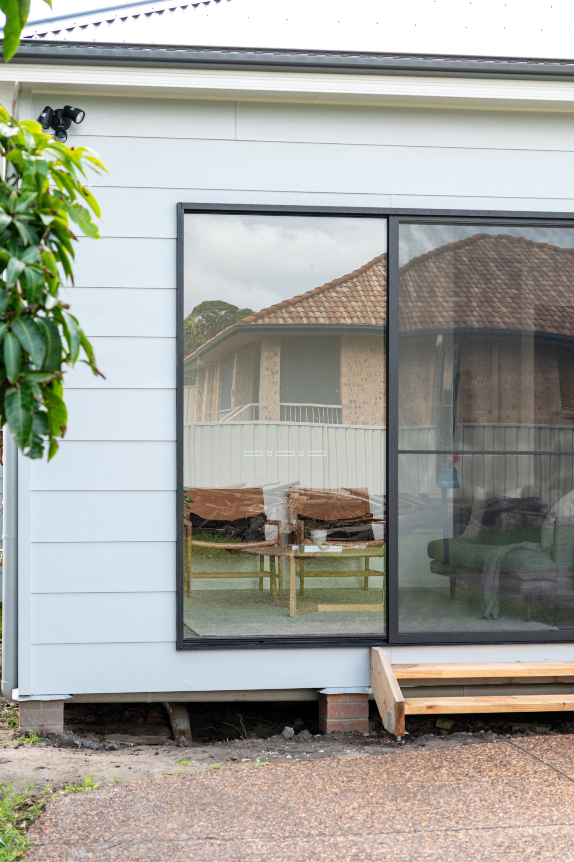 A window reflecting a brick house. Two chairs are visible. The building is light blue.