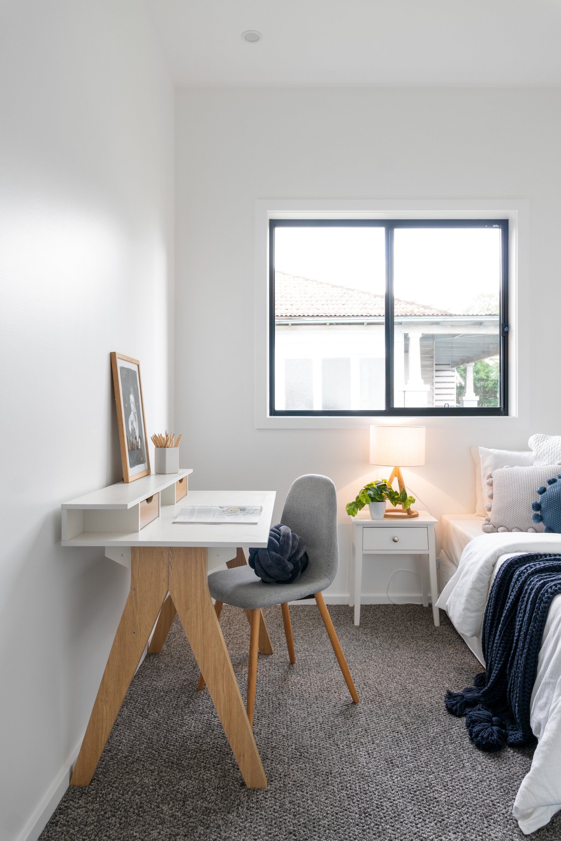 Bedroom with desk, chair, and bed next to a window. White walls, grey carpet, and natural wood accents.