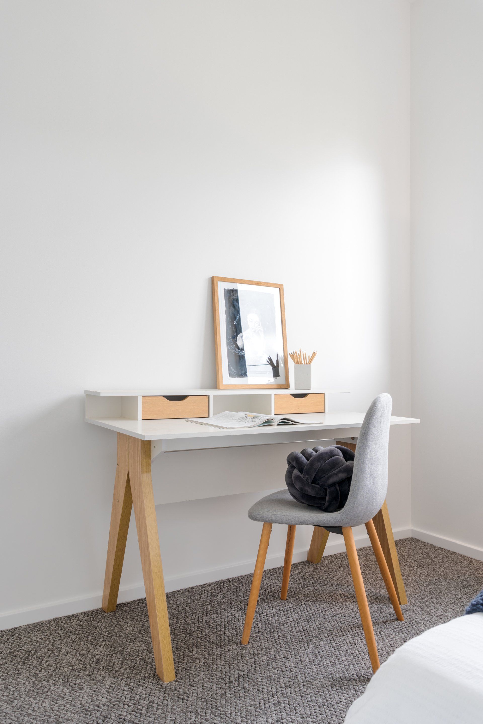 Desk with chair in a corner. White desk with wooden legs. Gray chair and carpet. Frame and pencil holder on desk.