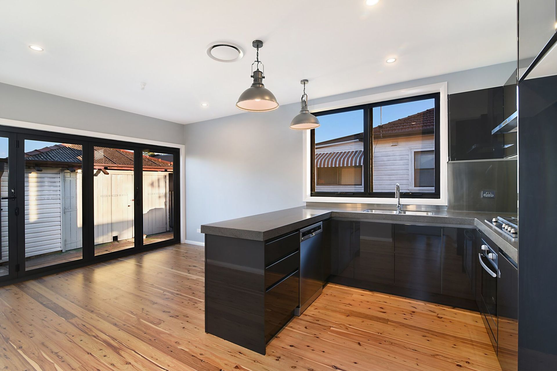 Modern kitchen with black cabinets, stainless steel appliances, and wood floors. Large glass doors open to a backyard.