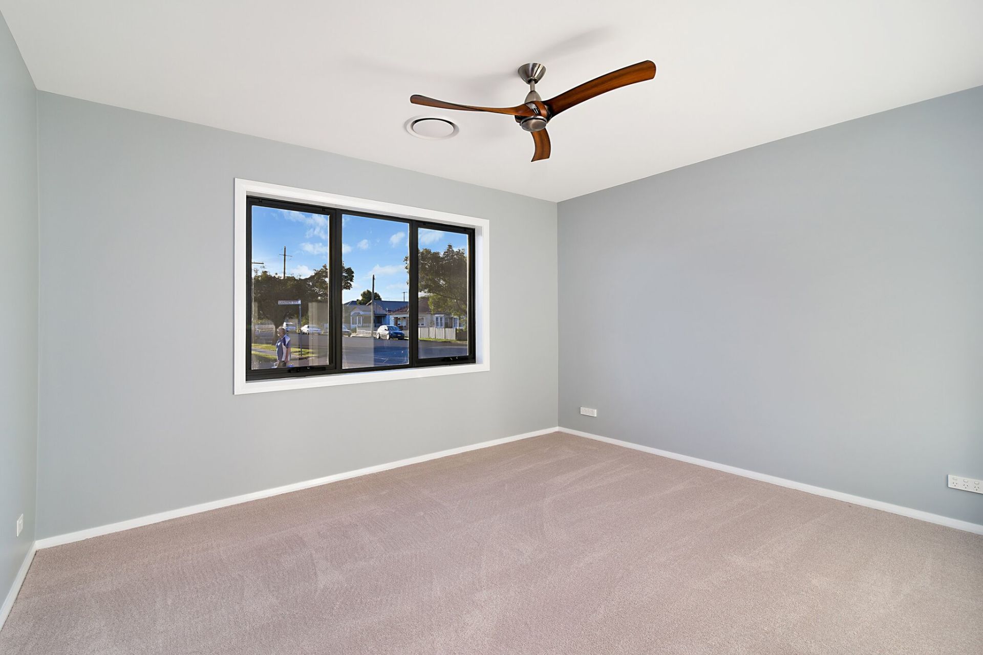 Empty bedroom with light blue walls, carpet, and a window with a view of the outside. A ceiling fan is visible.