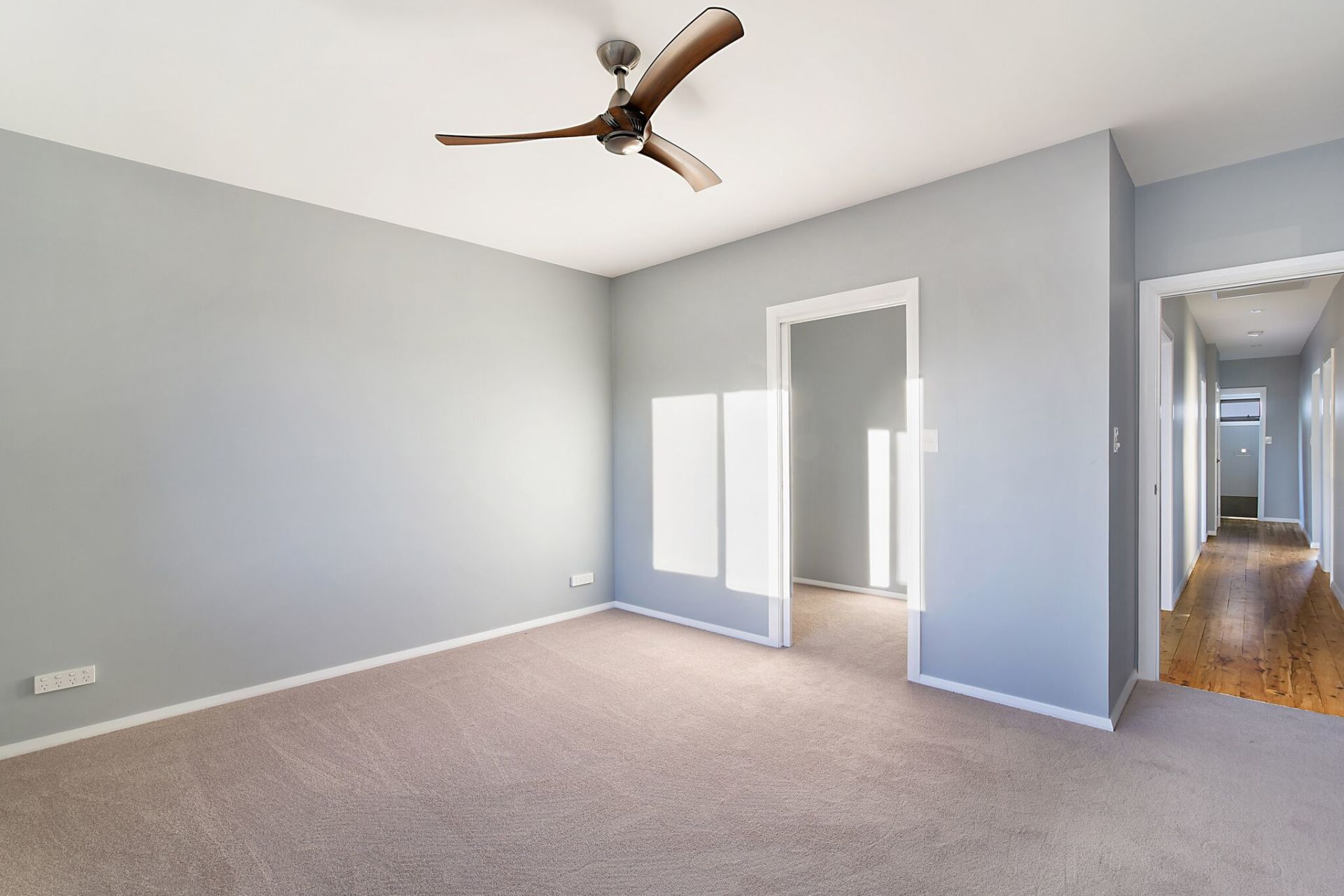 Empty room with light blue walls, tan carpet, and a ceiling fan. Hallway visible.