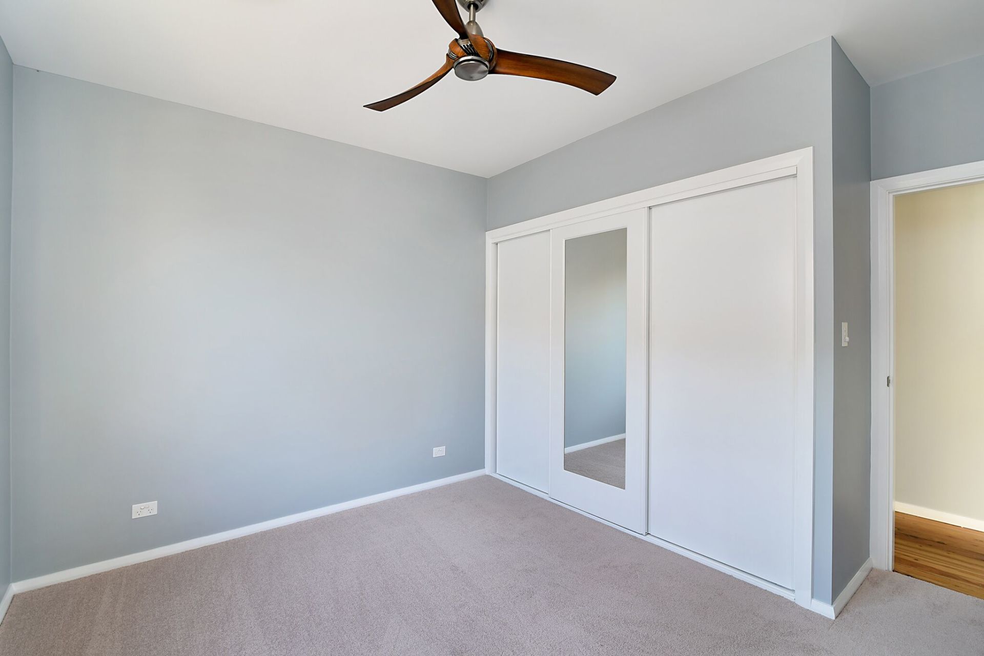 Empty bedroom with light gray walls, tan carpet, white closet doors, and a ceiling fan.