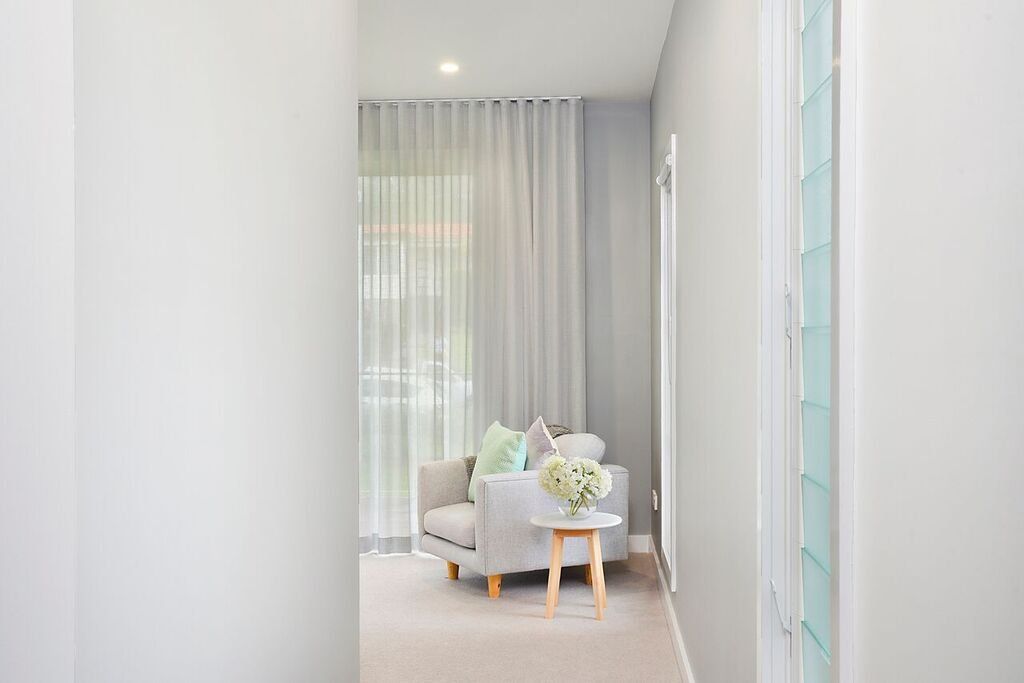 Narrow hallway leading to a light-filled sitting area with gray chair, sheer curtains, and small table.