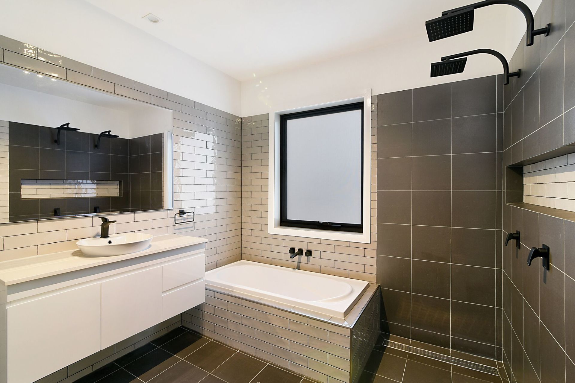 Modern bathroom with gray tile, white vanity, tub, and black fixtures.