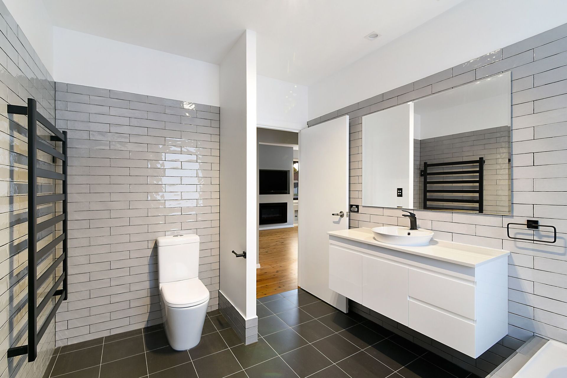 Modern bathroom with white vanity, toilet, and gray tiled walls; black towel warmer.