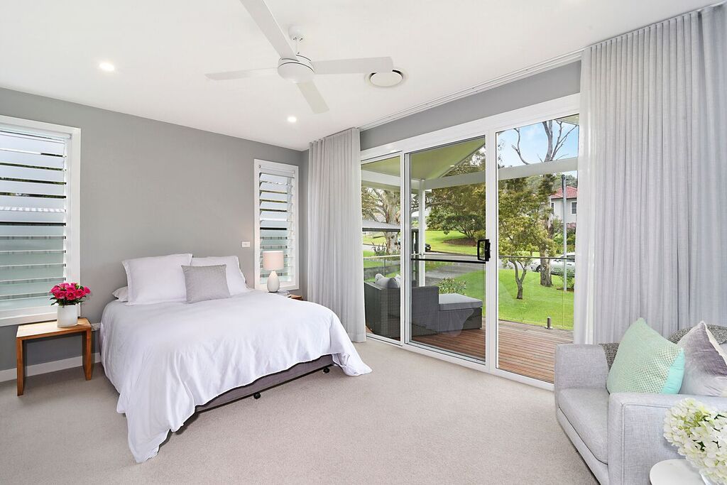 Bedroom with a bed, sofa, sliding glass doors, and a view of greenery. Gray walls, white bedding, and light-colored curtains.