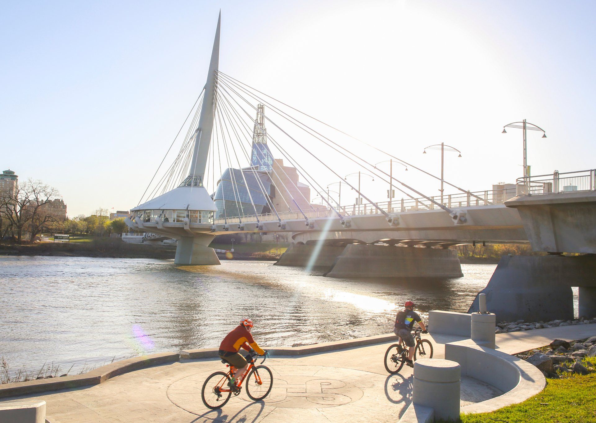 Cyclists near bridge with modern architecture over river, sunny day.