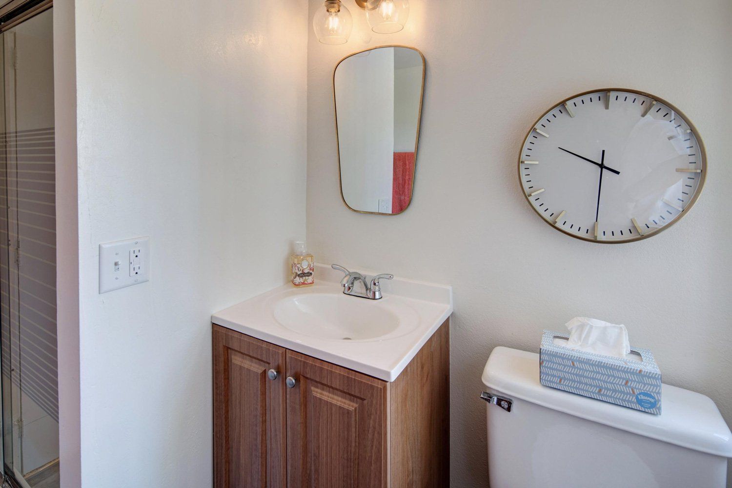 A bathroom with a sink , toilet , mirror and clock on the wall.
