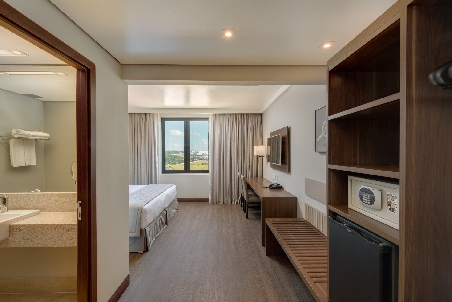 Quarto de hotel moderno com cama, escrivaninha, banheiro aberto e janela ampla com vista para a cidade.