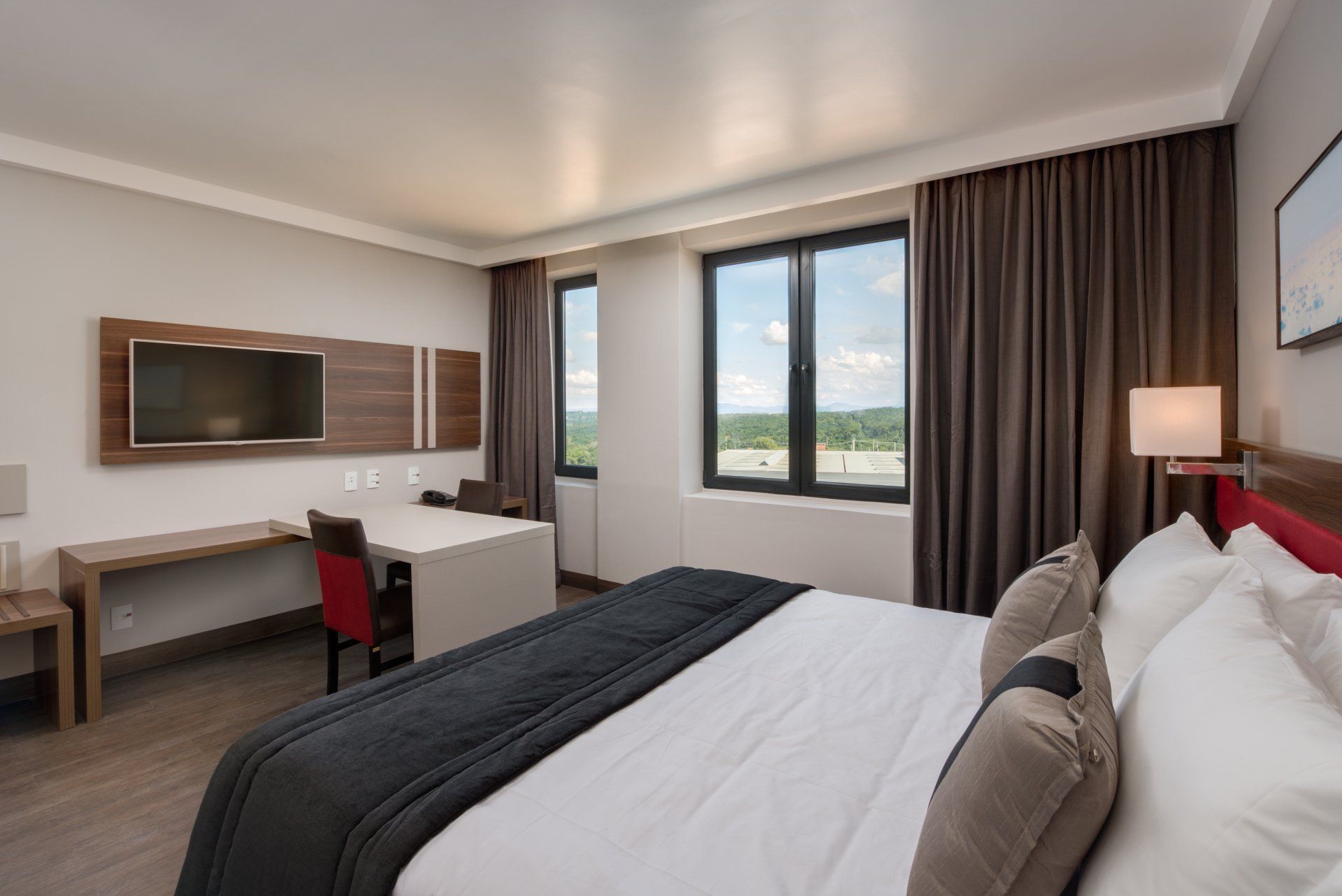 Quarto de hotel moderno com cama king-size, escrivaninha, TV de parede e janelas amplas com cortinas.