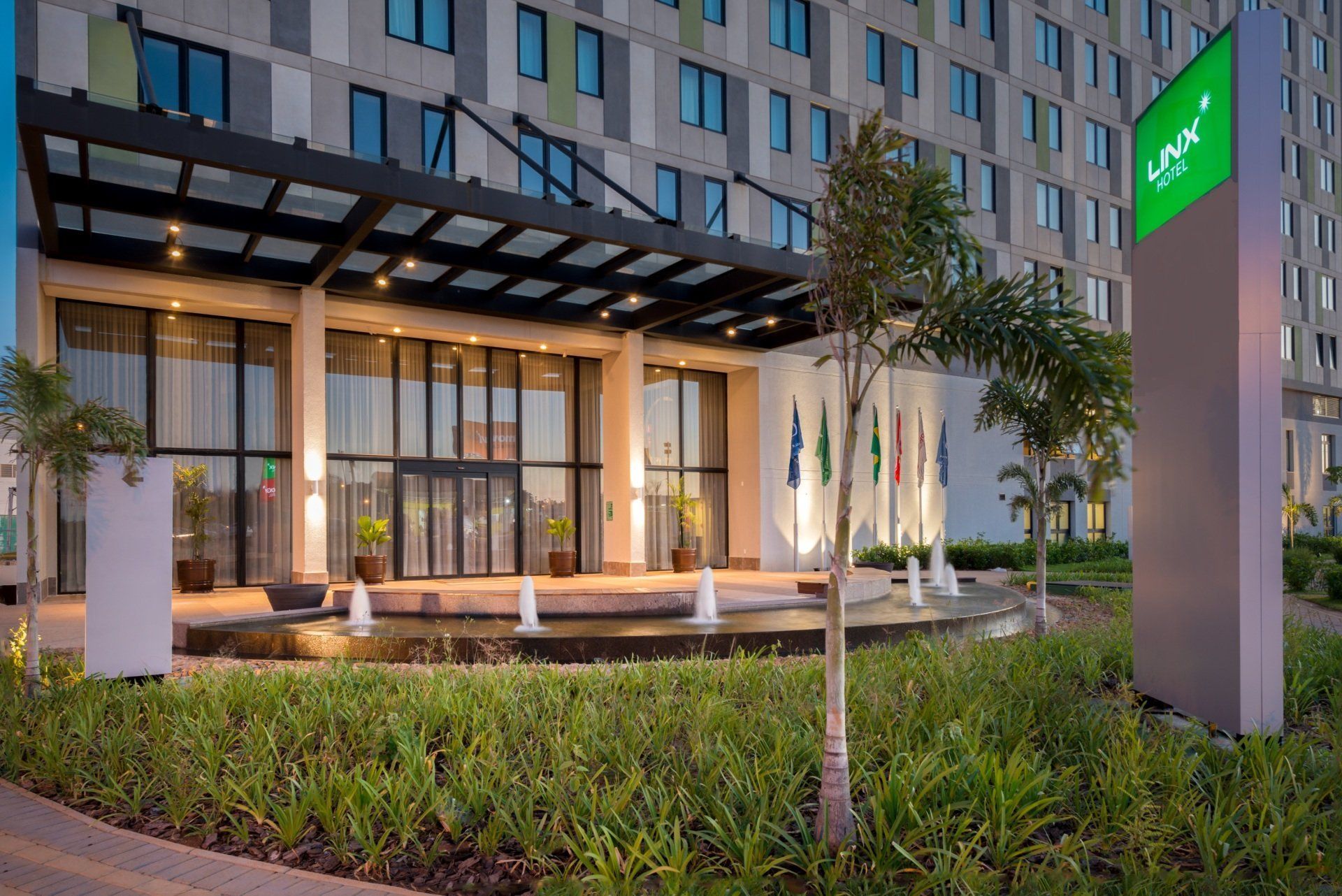 Entrada moderna do hotel com cobertura de vidro, passarela iluminada, árvores e uma placa verde ao entardecer.