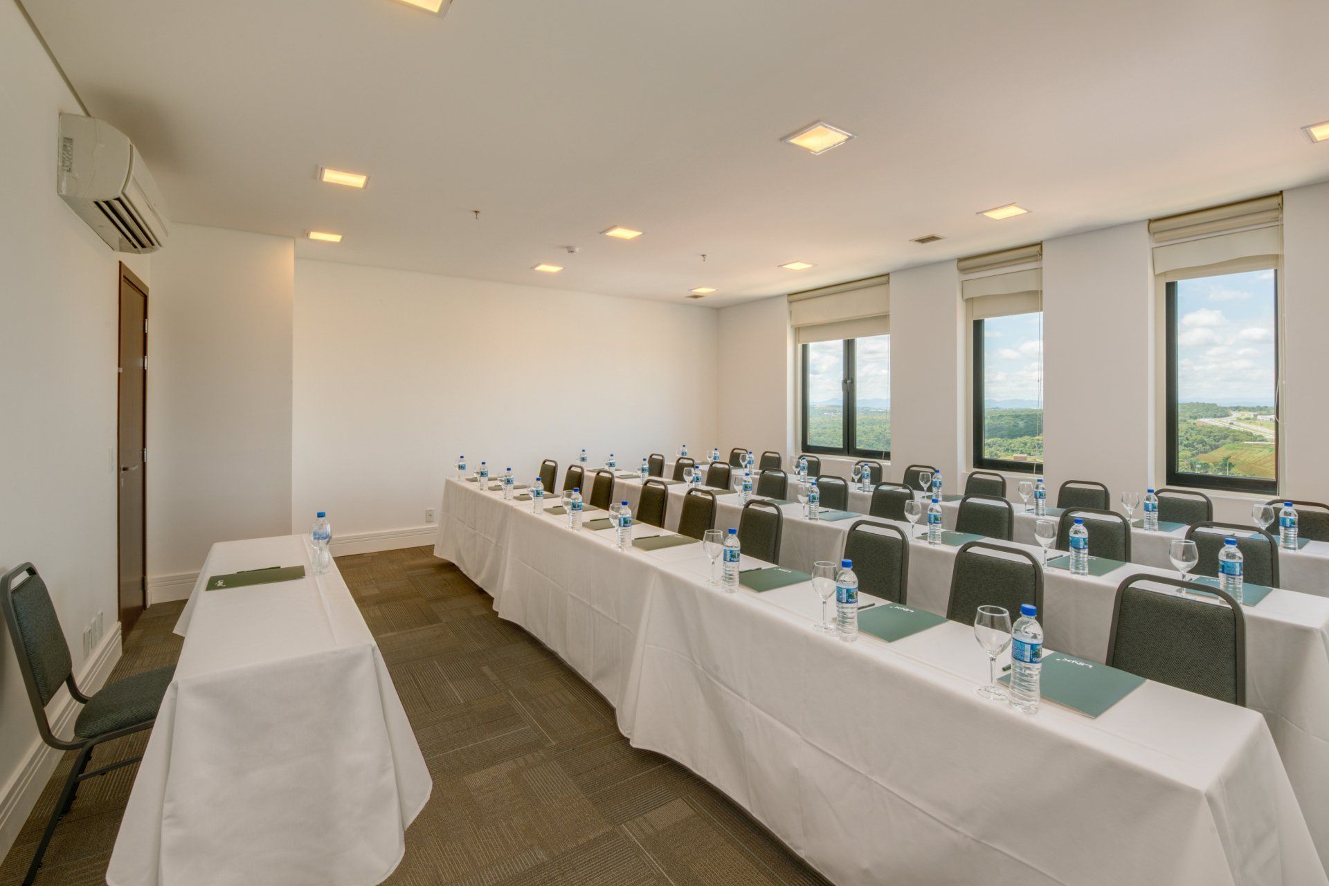 Sala de conferências vazia com mesas dispostas em formato de U, toalhas de mesa brancas, cadeiras e grandes janelas com vista para a cidade.