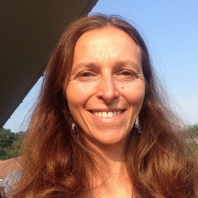 Una persona sorridente con lunghi capelli castano chiaro posa all'aperto contro un cielo azzurro limpido.