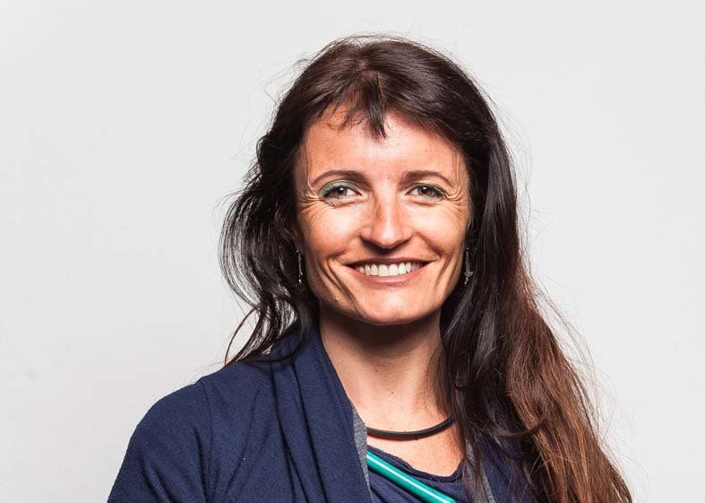 Una persona sorridente con lunghi capelli scuri, che indossa una maglietta blu scuro e un cordino verde, posa davanti a uno sfondo grigio uniforme.