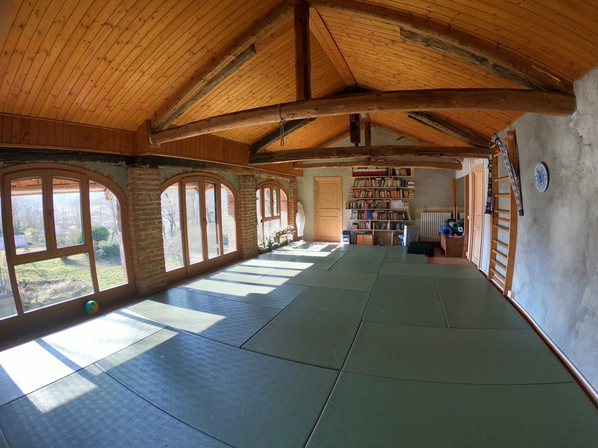 Un dojo al coperto con tatami verdi, soffitto rivestito in legno, travi a vista e finestre ad arco che si affacciano sulla natura.