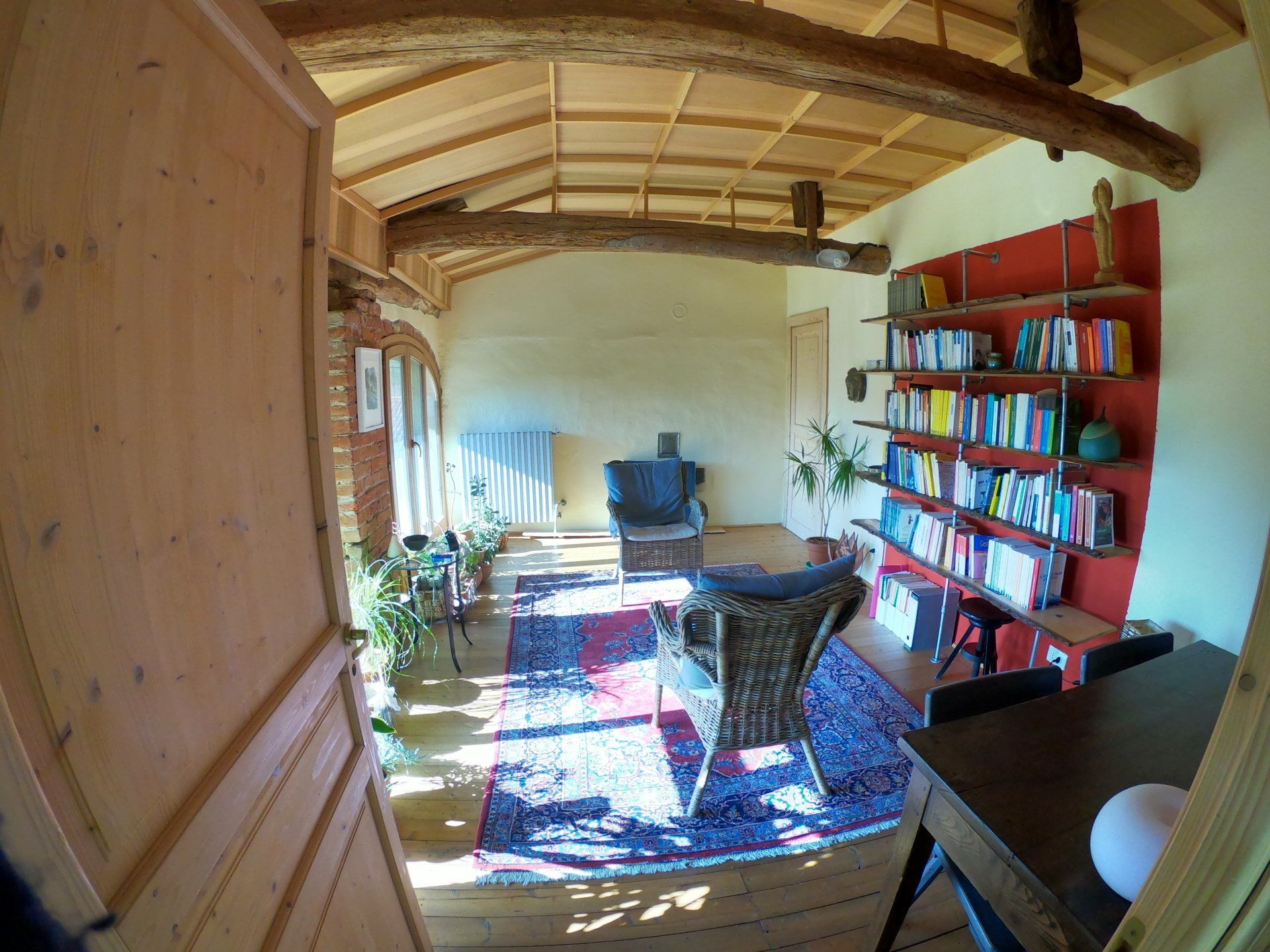 Una stanza luminosa con travi a vista sul soffitto, un tappeto colorato, due poltrone, una grande libreria contro una parete rossa e una scrivania.