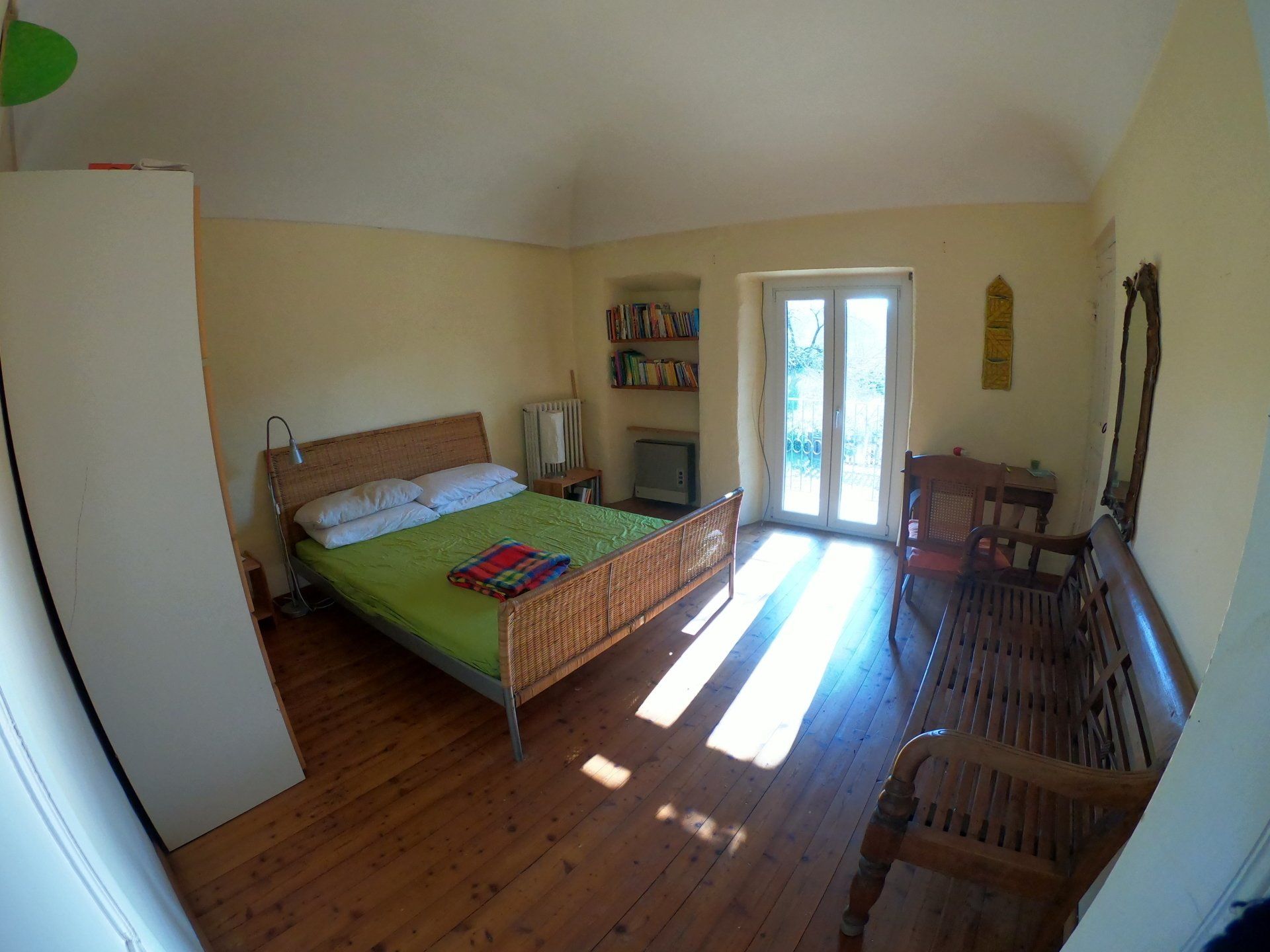Una camera da letto con pareti gialle e pavimento in legno, arredata con un letto con copriletto verde, un armadio e una panca in legno.