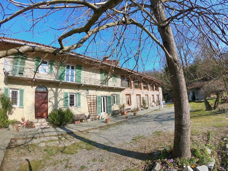Una casa in pietra color sabbia a due piani con persiane verdi e un cortile di ghiaia sotto un cielo azzurro e limpido.