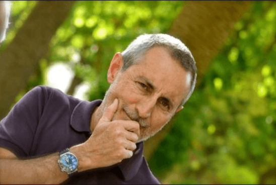 Una persona con una polo viola e un orologio da polso si sporge verso la telecamera, appoggiando il mento sulla mano su uno sfondo verde.