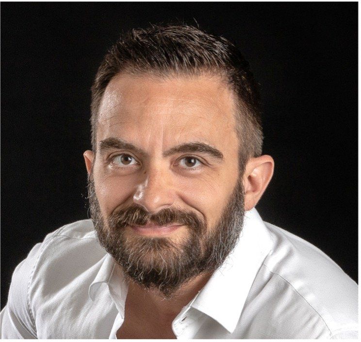 Un uomo con la barba e una camicia bianca sorride su uno sfondo nero.