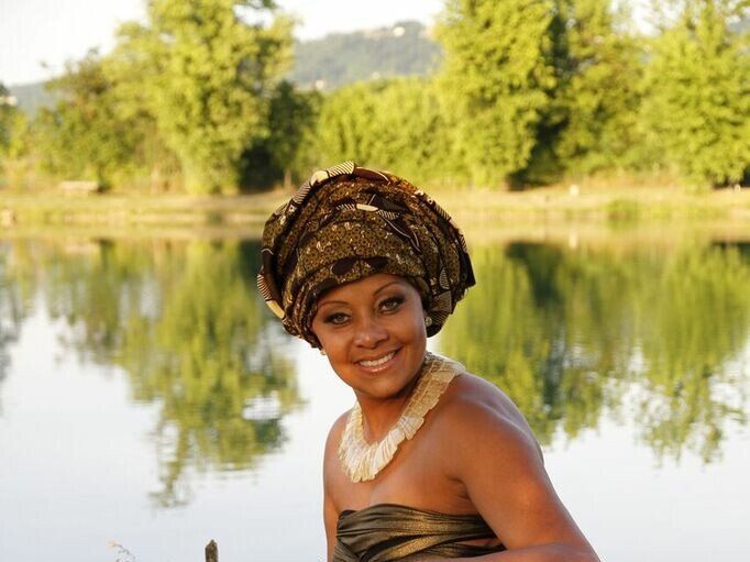 Una persona sorridente indossa un elaborato turbante scuro e una collana a più fili di pietre chiare, all'aperto in riva a un lago tranquillo.