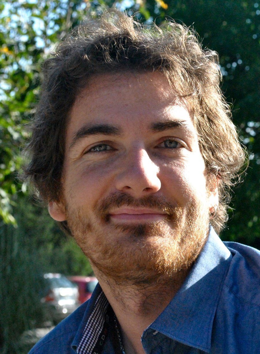 Primo piano del volto di una persona con capelli scuri e ricci, occhi chiari e una leggera barba incolta, che sorride dolcemente su uno sfondo sfocato.
