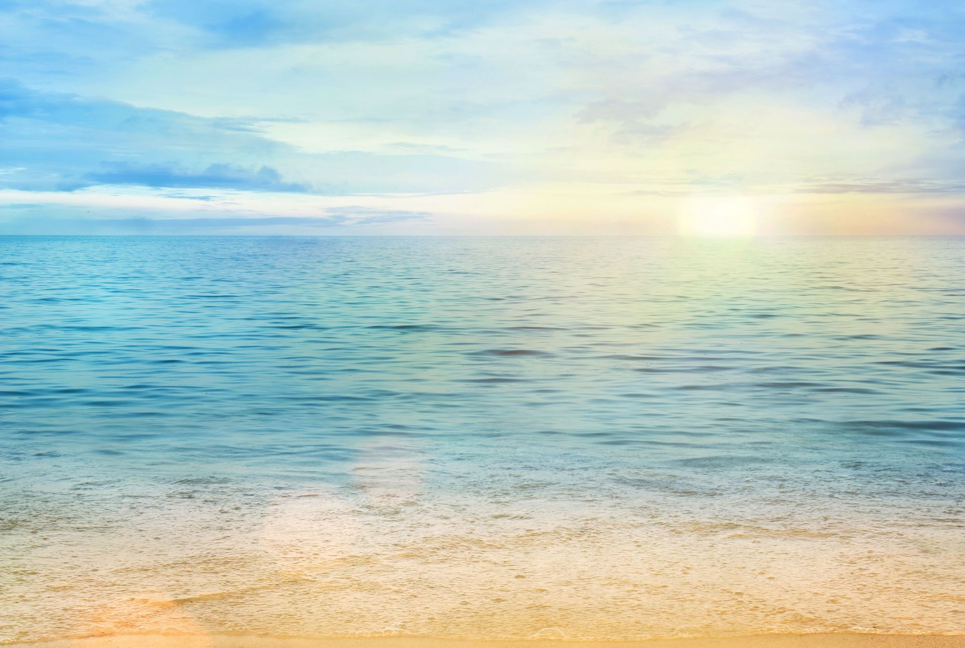 Una scena di spiaggia illuminata dal sole, con una riva sabbiosa, dolci onde azzurre e un cielo velato e dai toni tenui al tramonto.