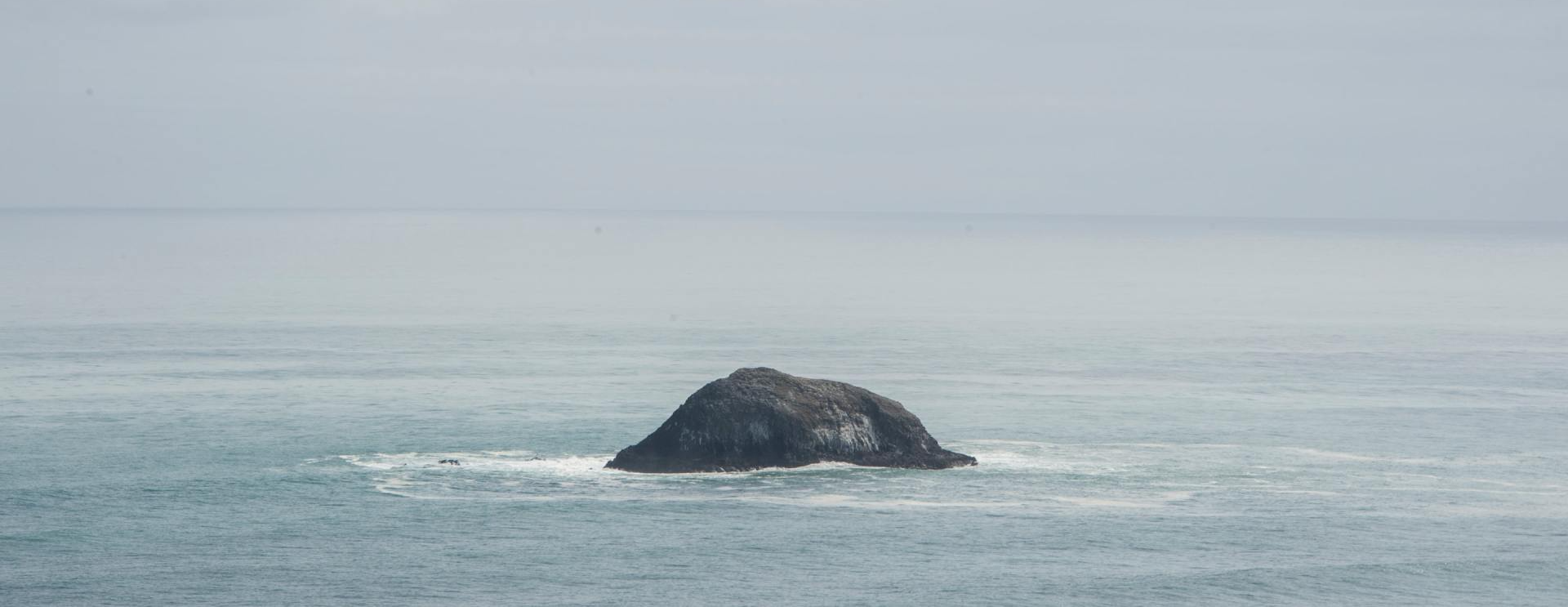 Un'unica isola scura e rocciosa si erge al centro di un oceano calmo e immenso, sotto un cielo nebbioso e nuvoloso.