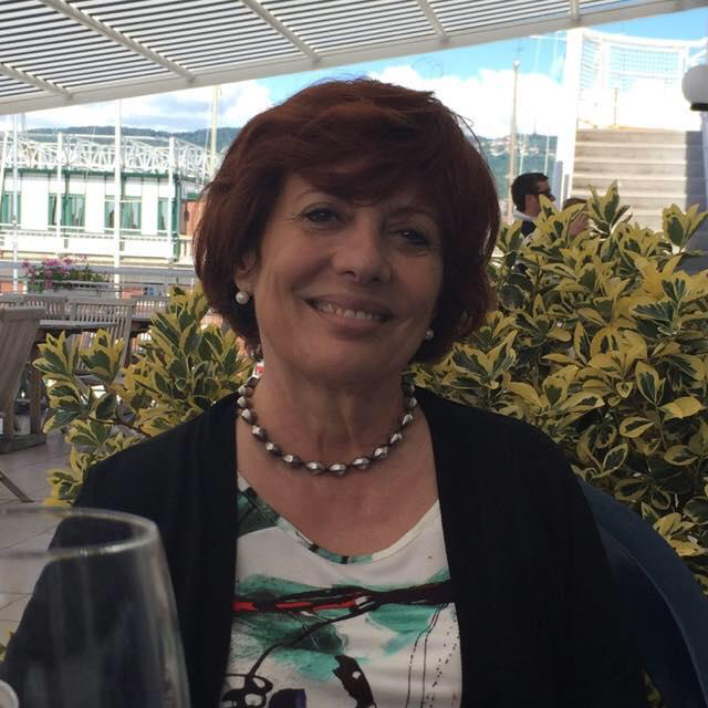 Una donna dai capelli ramati sorride mentre indossa una collana di perline e una blusa a fantasia, seduta in un ristorante all'aperto.